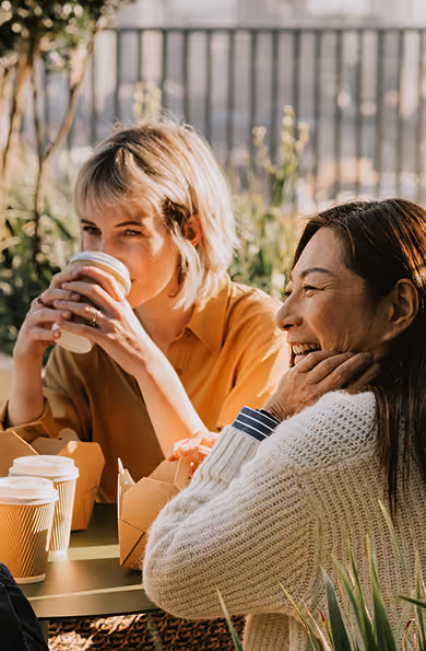 Two women sitting outdoors at a table with coffee and takeout boxes, one woman smiling and the other drinking from a paper cup.