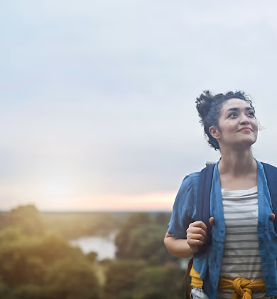 A young woman with curly hair tied up, wearing a striped shirt and blue jacket with a yellow sweater tied around her waist, stands outdoors with a backpack, looking up and smiling slightly against a backdrop of a river and green landscape at sunset.