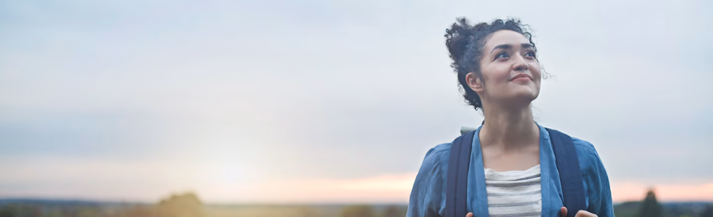 Young woman with curly hair tied up wearing a backpack and a blue jacket, looking slightly up and to the side with a calm expression against a soft, cloudy sky at sunset.