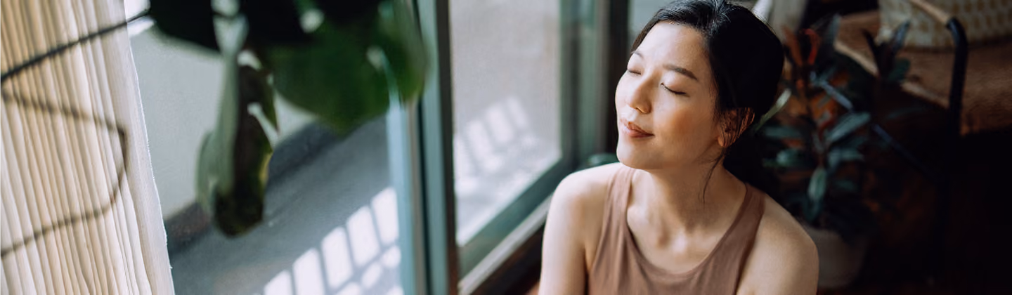 A serene young Asian woman with closed eyes sitting near large windows, bathed in natural light with green plants and cozy furniture.