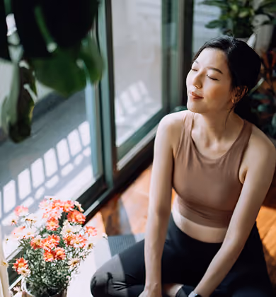 A woman sitting cross-legged on the floor near a window with sunlight, eyes closed, enjoying a peaceful moment beside a pot of colorful flowers.