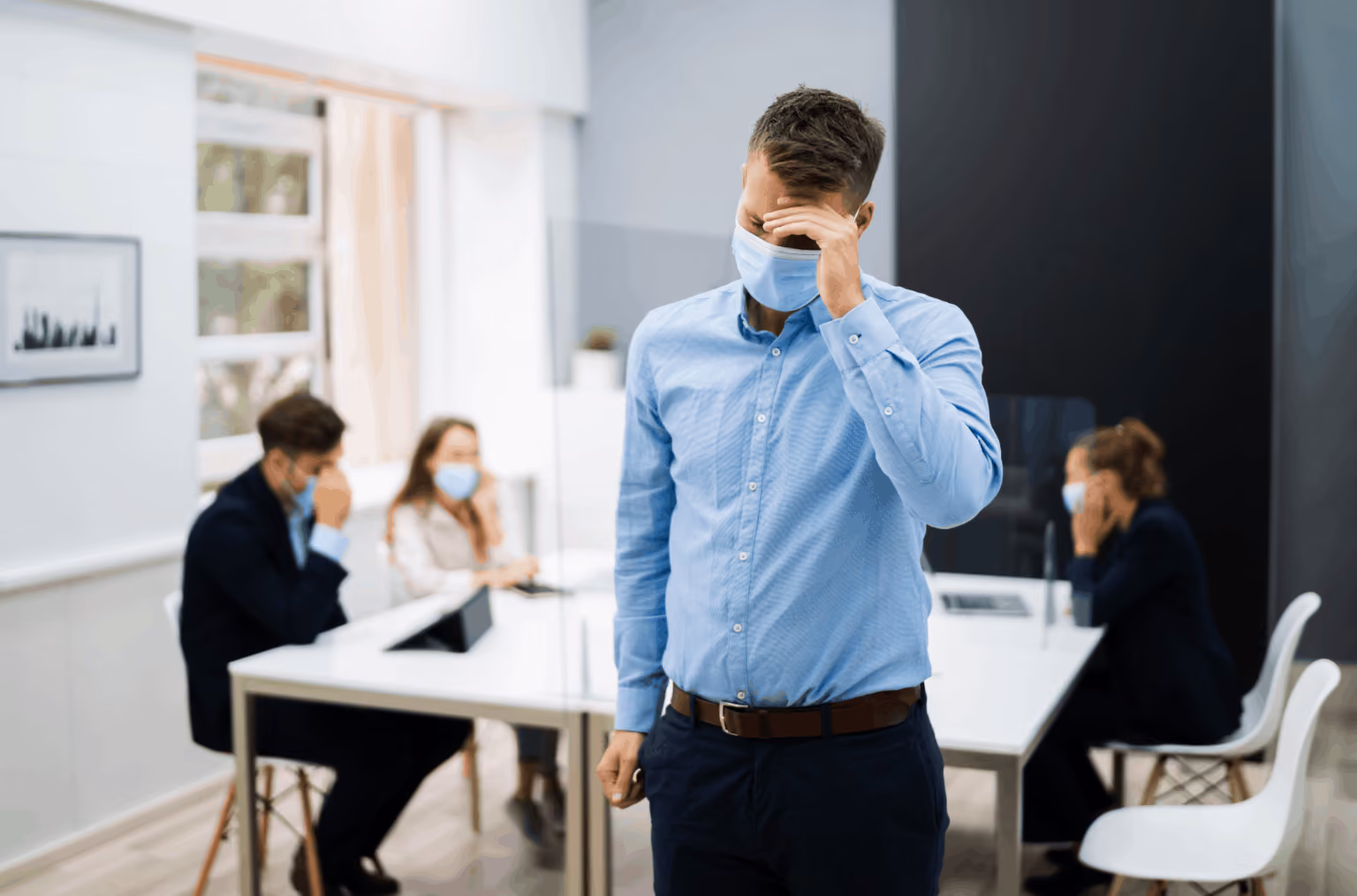 Man in a blue shirt and blue face mask with his hand on his face in the office
