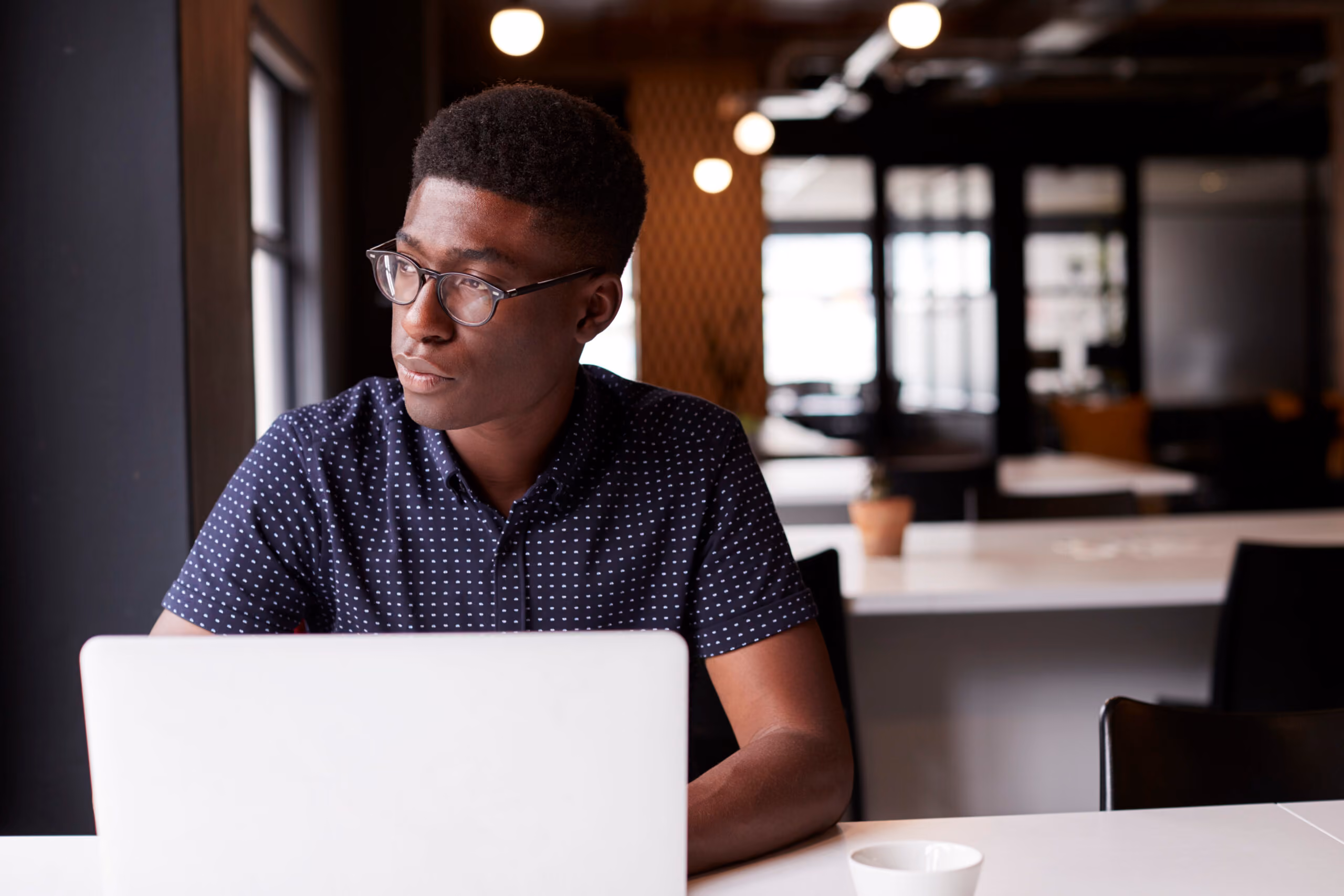 An anxious man with glasses on looking out a window while on his laptop