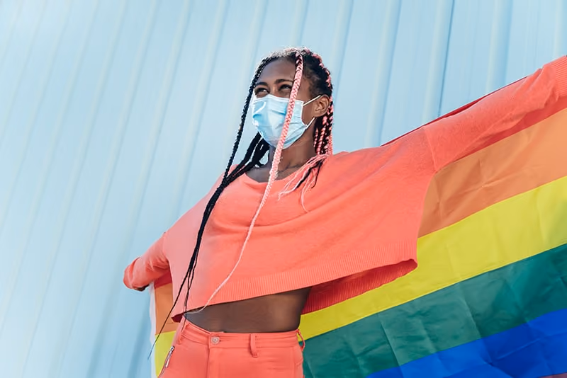 Woman in a facemask holding a rainbow flag behind her back
