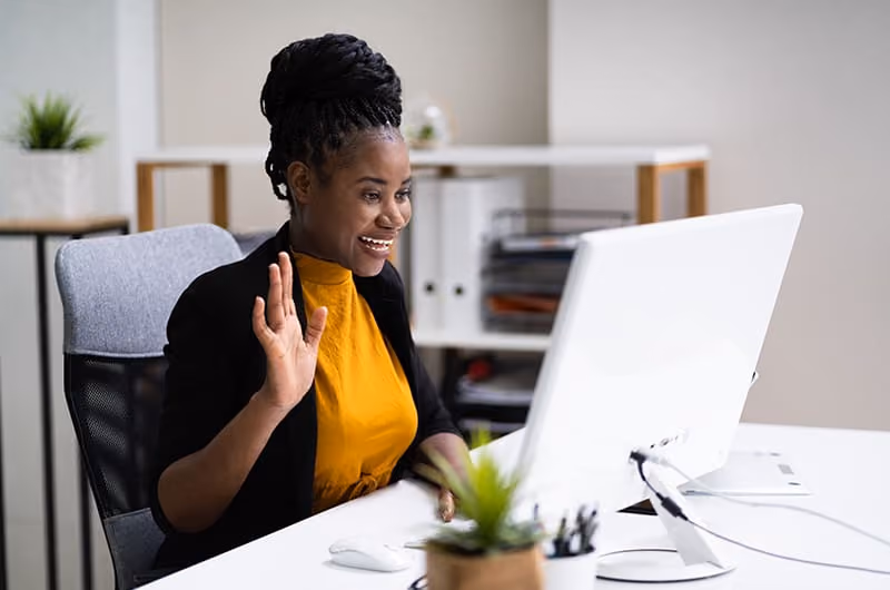Woman in an orange shirt and a black cardigan waving to someone on a laptop