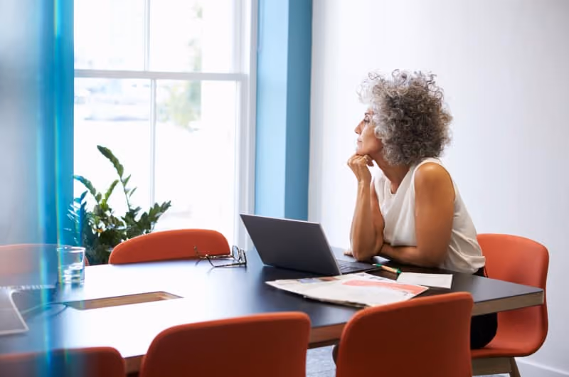 A woman sitting at a desk, looking out of a window while using her laptop