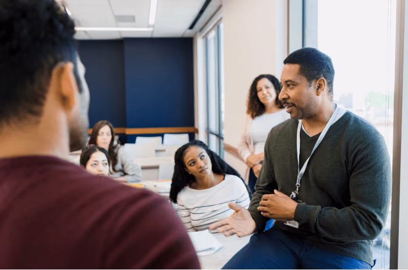 A Hispanic man with a green sweater and a lanyard speaking to a group of colleagues at his job 