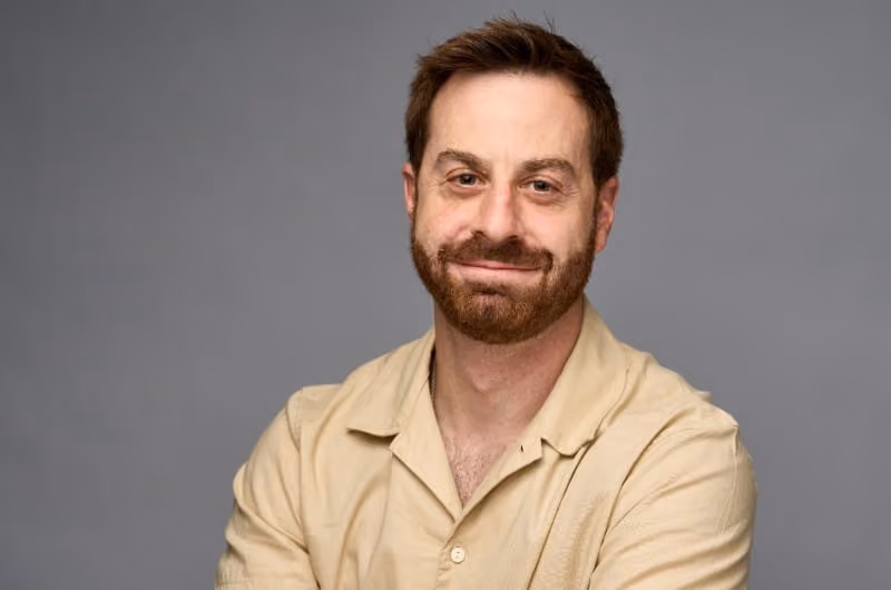 Eli Greenwald in a tan button down shirt in front of a grey backdrop