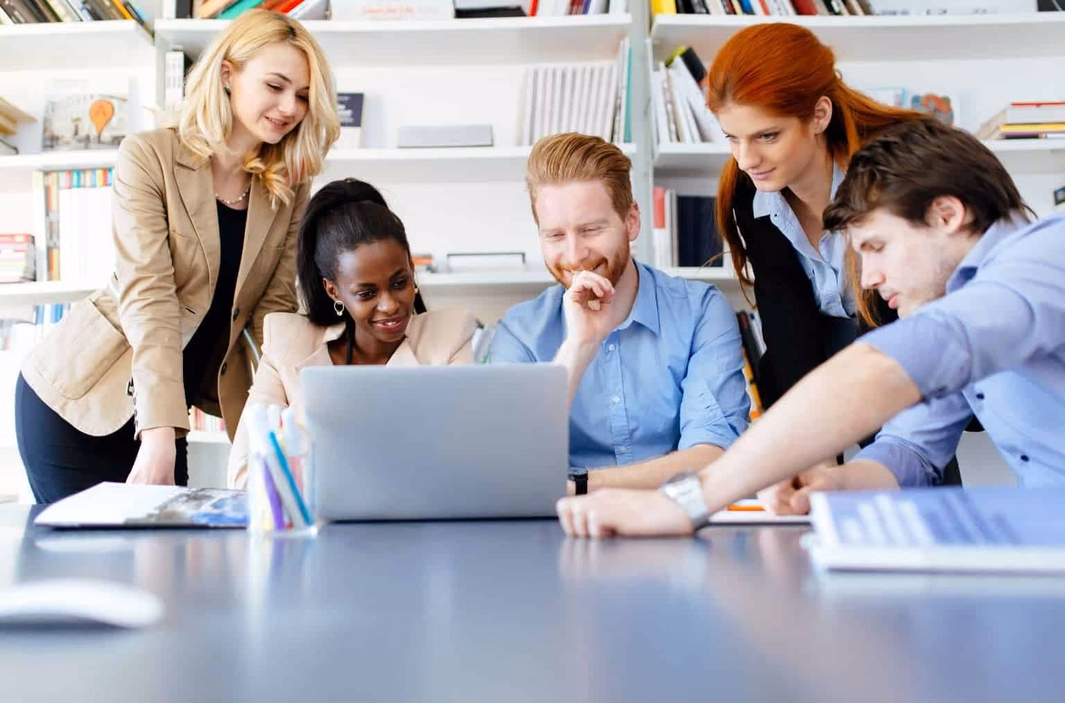 Coworkers around a man in a blue shirt looking at his laptop