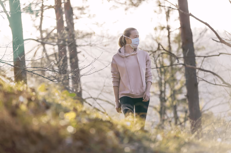 A woman in a face mask, pink hoodie and black leggings walking through the forest