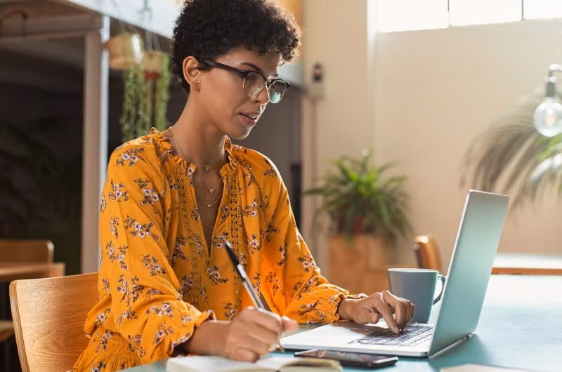 Woman with glasses in an orange floral shirt taking notes while working on a laptop