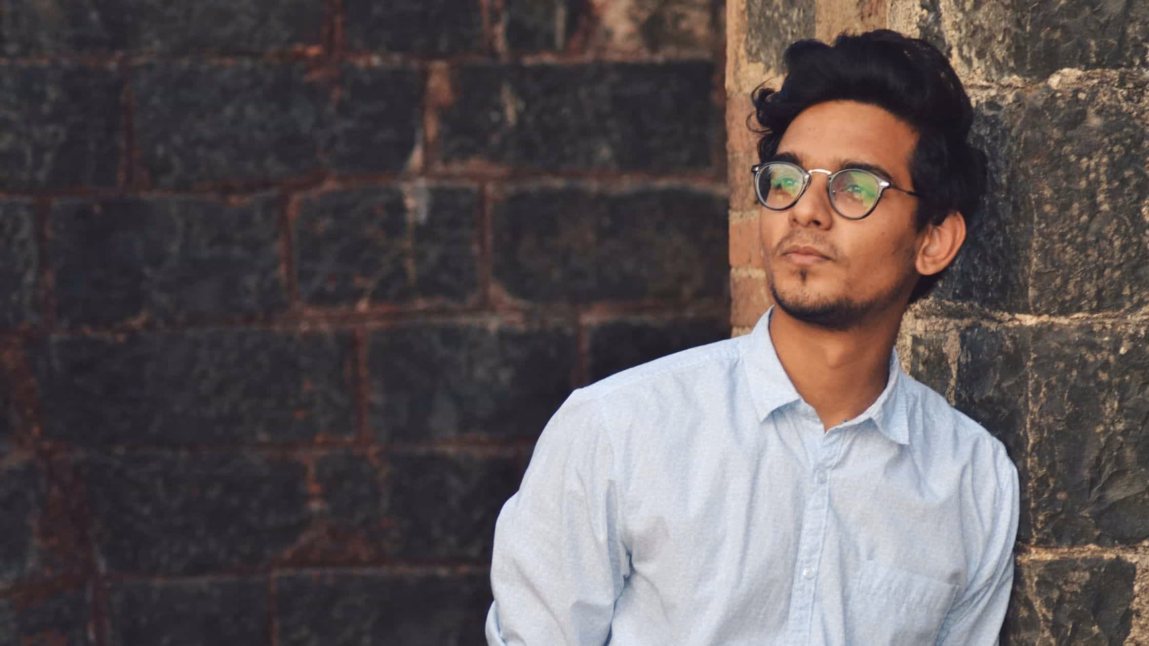 Man in a button shirt with glasses leaning on a brick wall