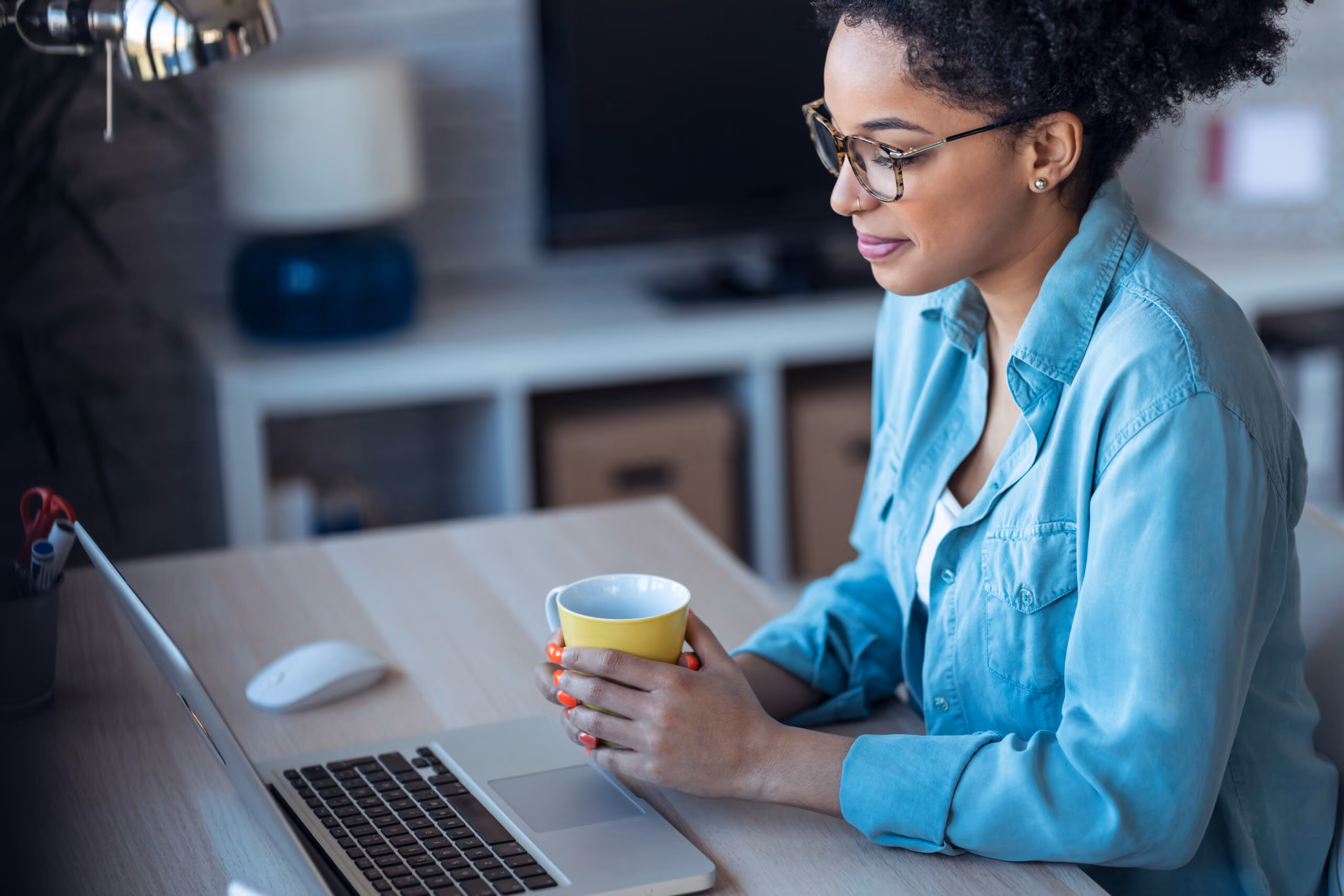 Woman with glasses and a nose piercing in a blue shirt holding a mug and looking at a laptop