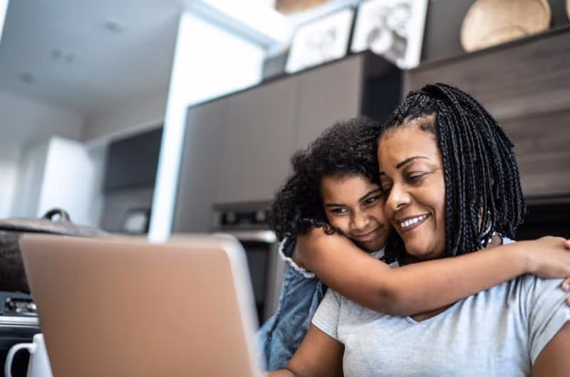 A woman looking at her laptop while her young daughter hugs her