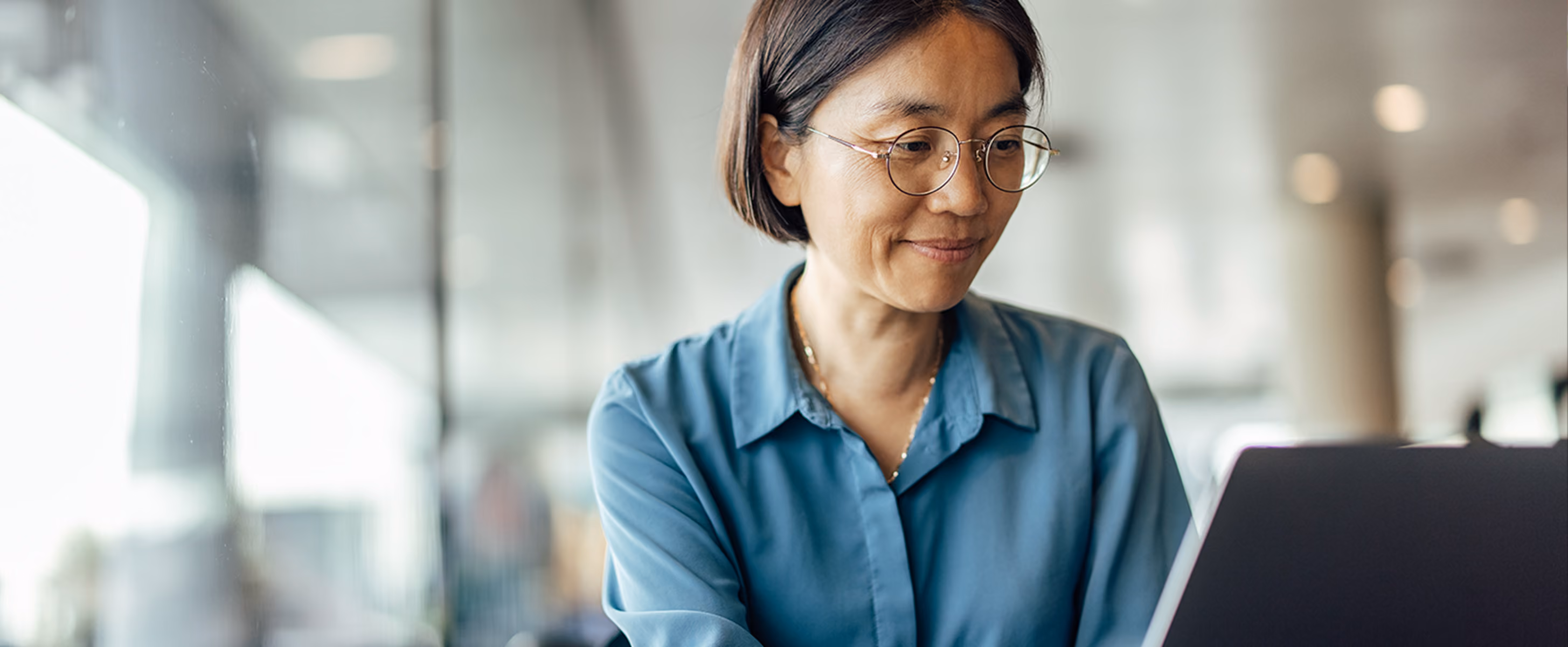 A woman in a blue shirt with round glasses working on a laptop