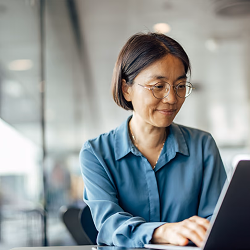 A woman in a blue shirt with round glasses working on a laptop