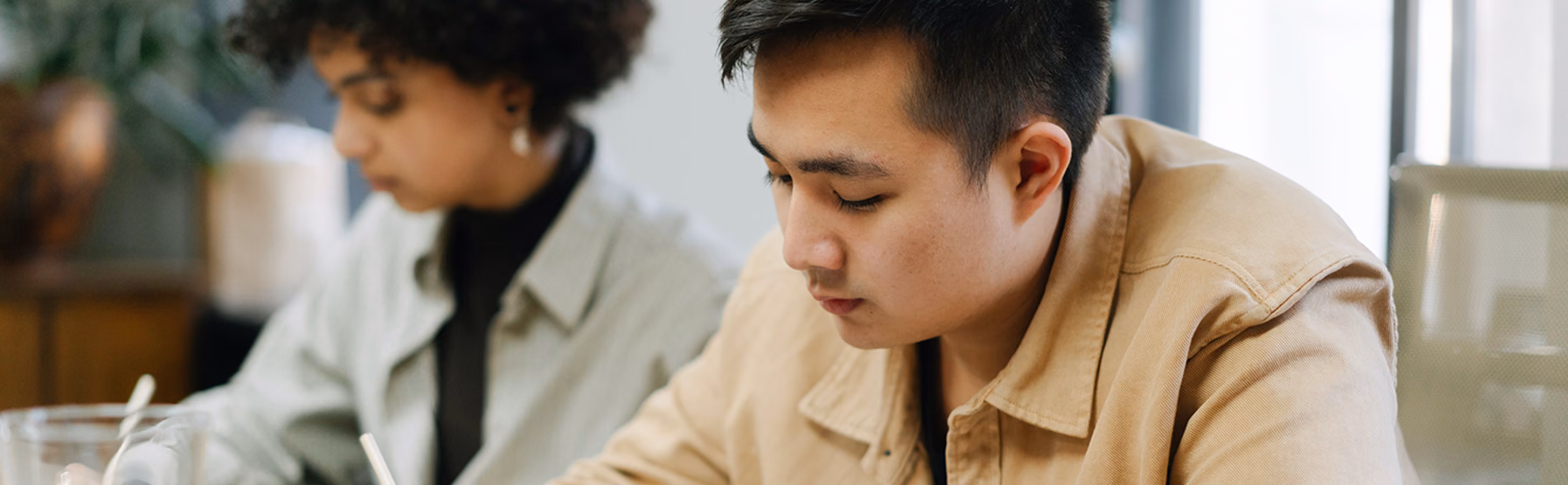 An Asian man in a tan jacket writing notes with a pen in a notebook