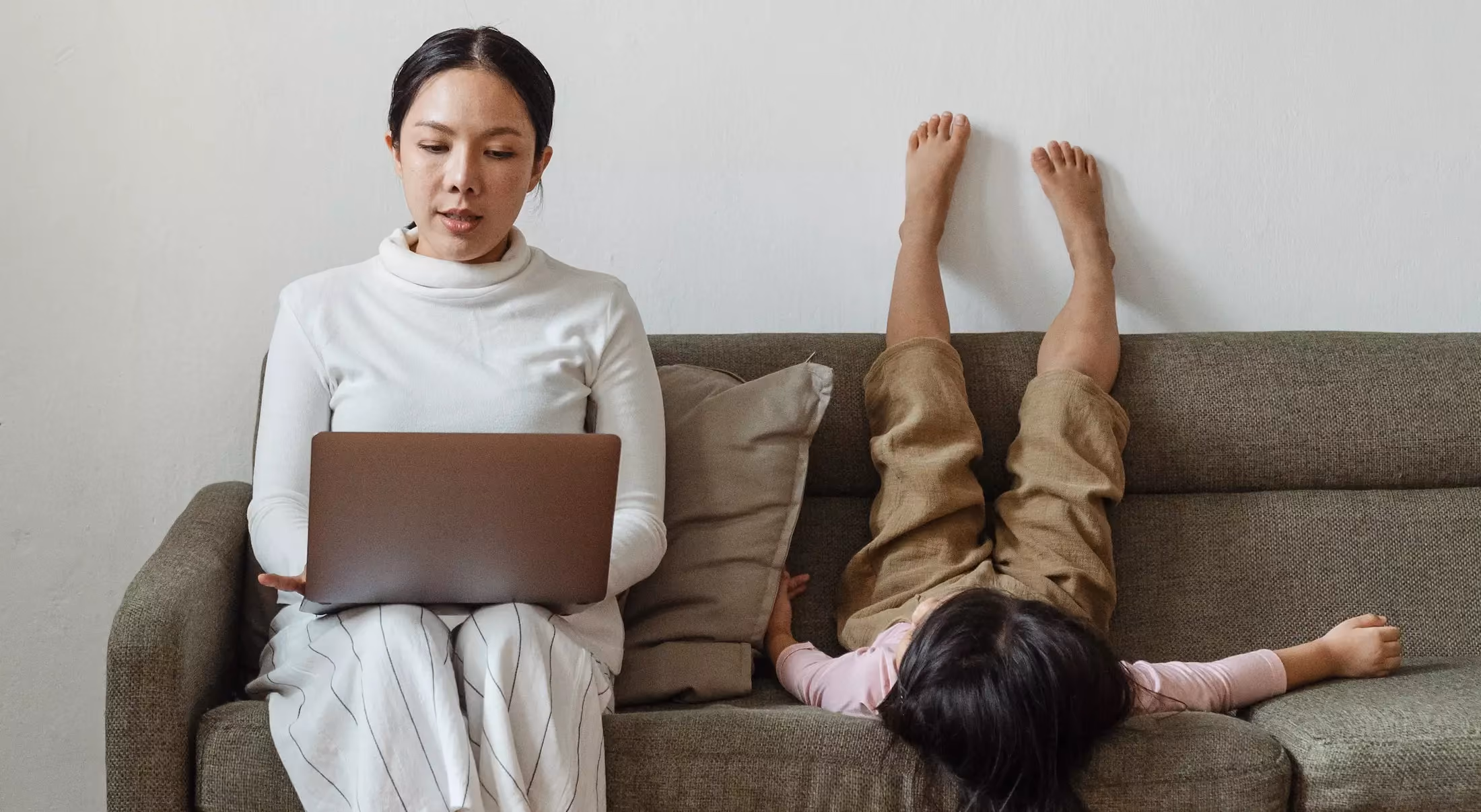 A woman in a white sweater working on a laptop on the couch while her child is upsidedown beside her