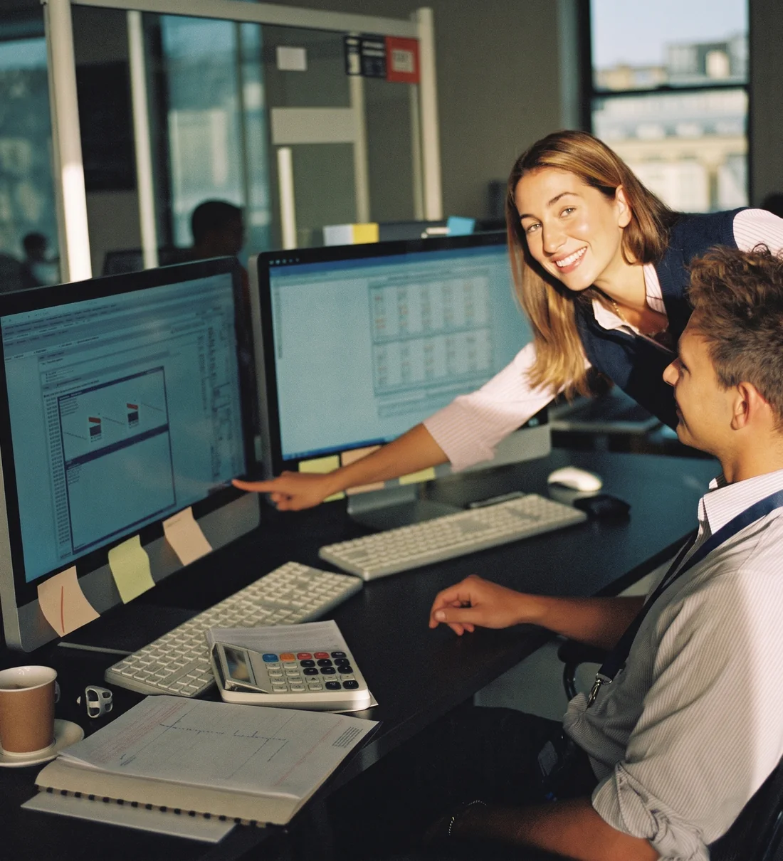 A senior manager leaning over a staff accountant's desk, pointing at a spreadsheet on his monitor
