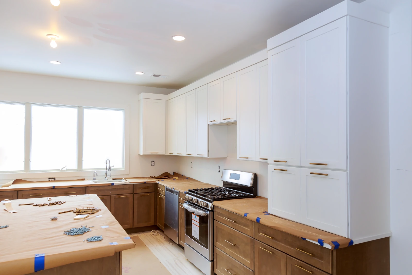 Kitchen cabinet painting project in progress showing white-painted upper cabinets alongside unpainted wood lower cabinets with countertops masked for protection