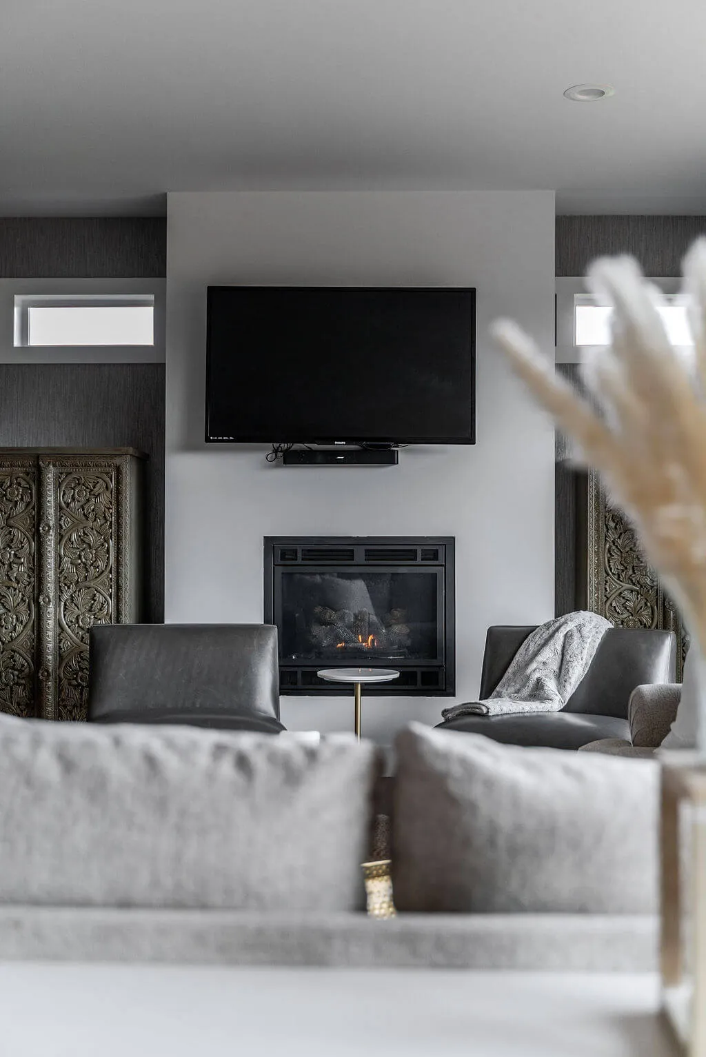 Modern living room with a wall-mounted flat screen TV above a lit gas fireplace, flanked by dark carved wooden panels, two gray leather chairs with a gray throw blanket on one, and a small round side table between the chairs.
