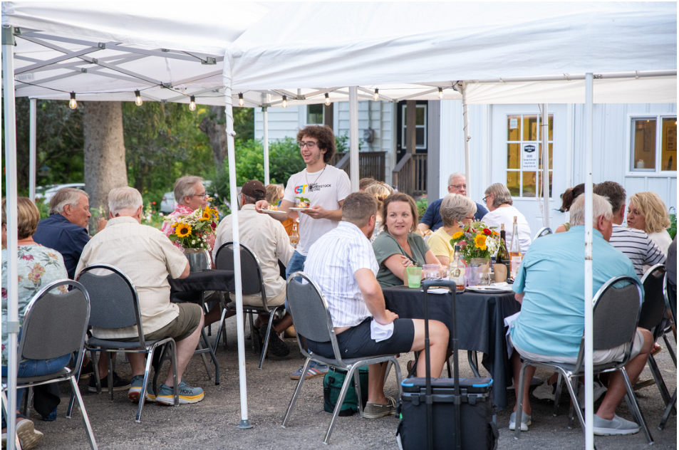 An outdoor dinner and live music event under white canopy tents on a sunny day. Guests sit at round tables, enjoying food and conversation. A young man plays a keyboard with sheet music in front of him, providing live entertainment. The event takes place on a paved area surrounded by lush green trees.