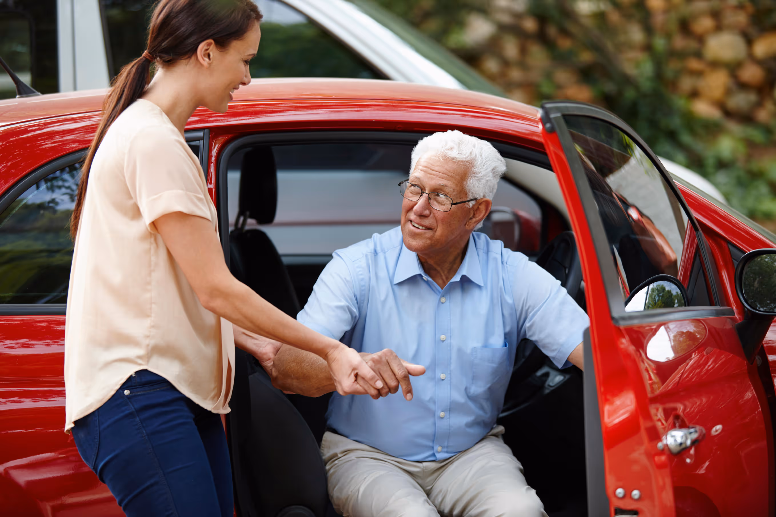 Caregiver assisting an older man getting out of a red car as part of an ambulatory transportation service