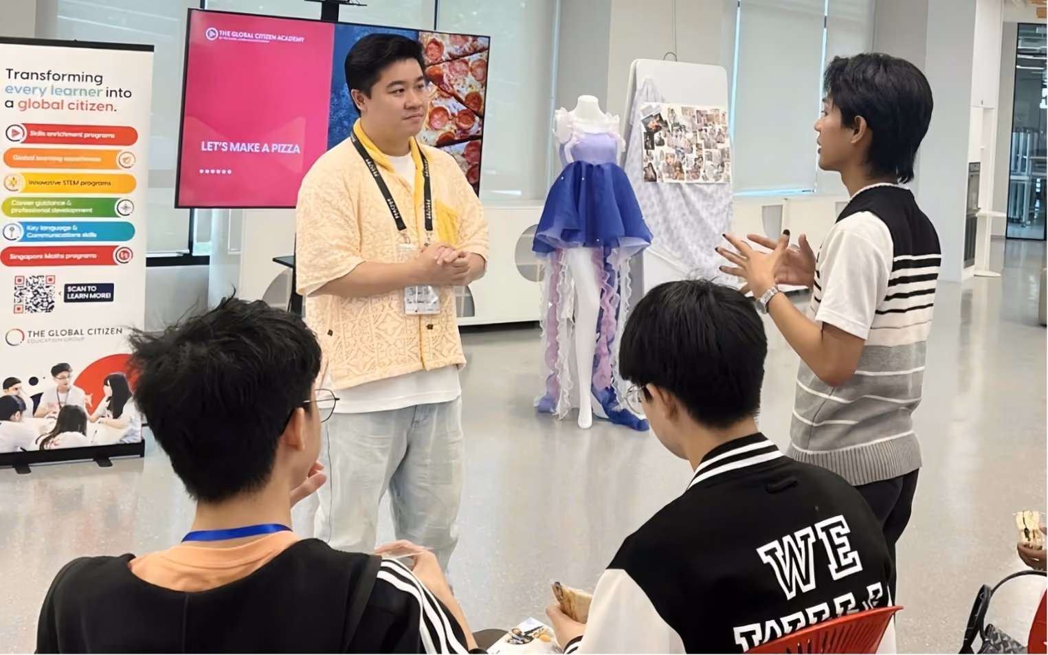 A man in a yellow shirt speaking to a small group of young people seated in a classroom with educational posters and a mannequin dressed in a blue and purple outfit in the background.