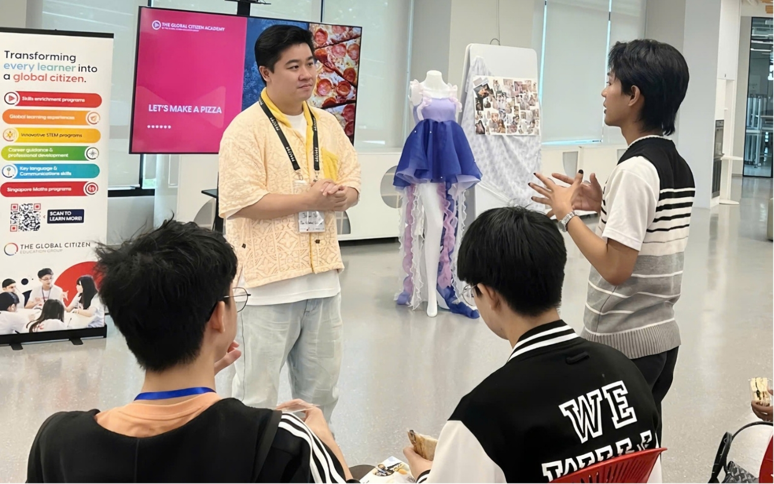 A group of young people engaged in a discussion in an indoor setting with a mannequin dressed in a purple and blue outfit and a screen displaying 'LET'S MAKE A PIZZA'.