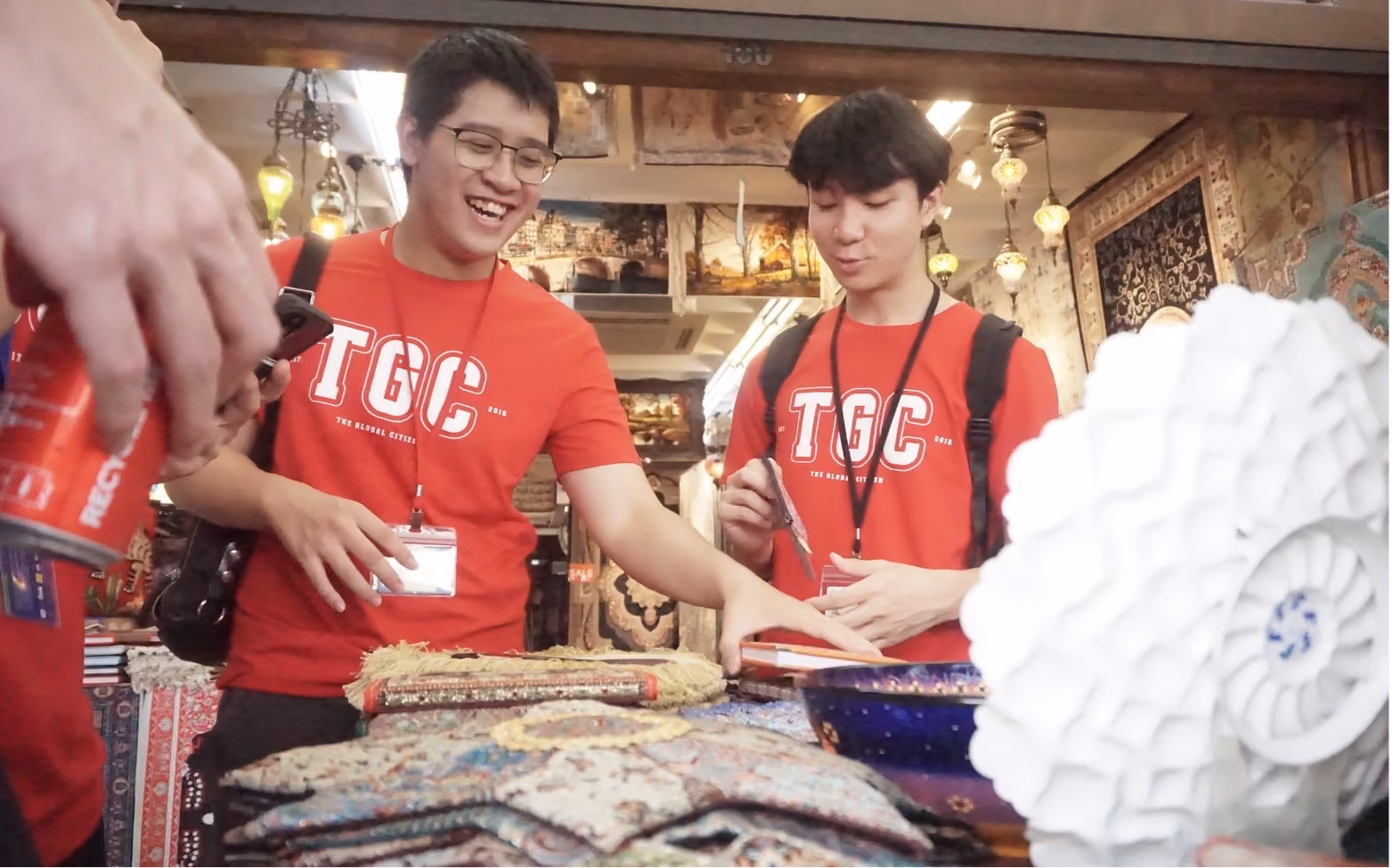 Two young men in red TGC shirts examining rugs and decorative items in a market stall.