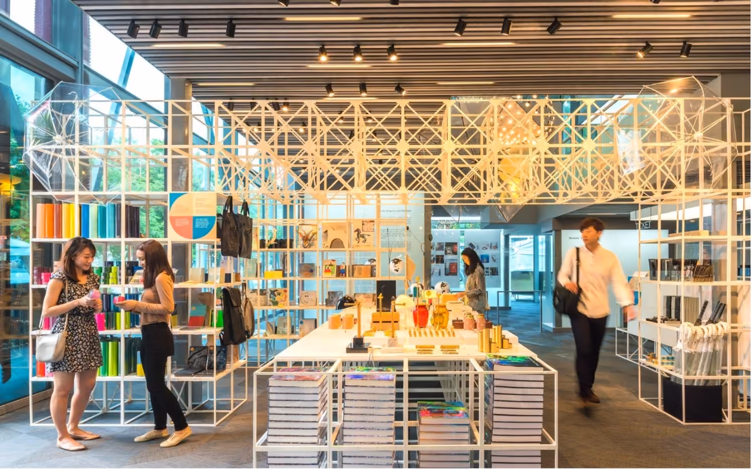 Modern retail store interior with white metal grid shelving displaying colorful items, books, umbrellas, and decorative objects, with three people browsing.