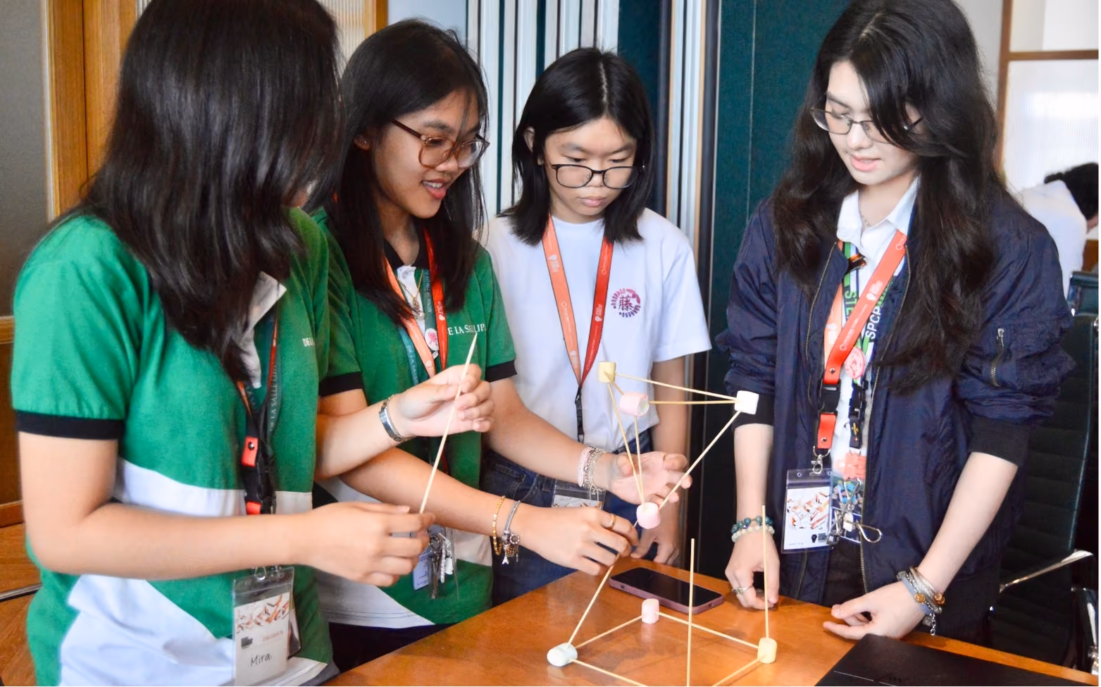 Four young women collaboratively building a structure with marshmallows and sticks at a wooden table.