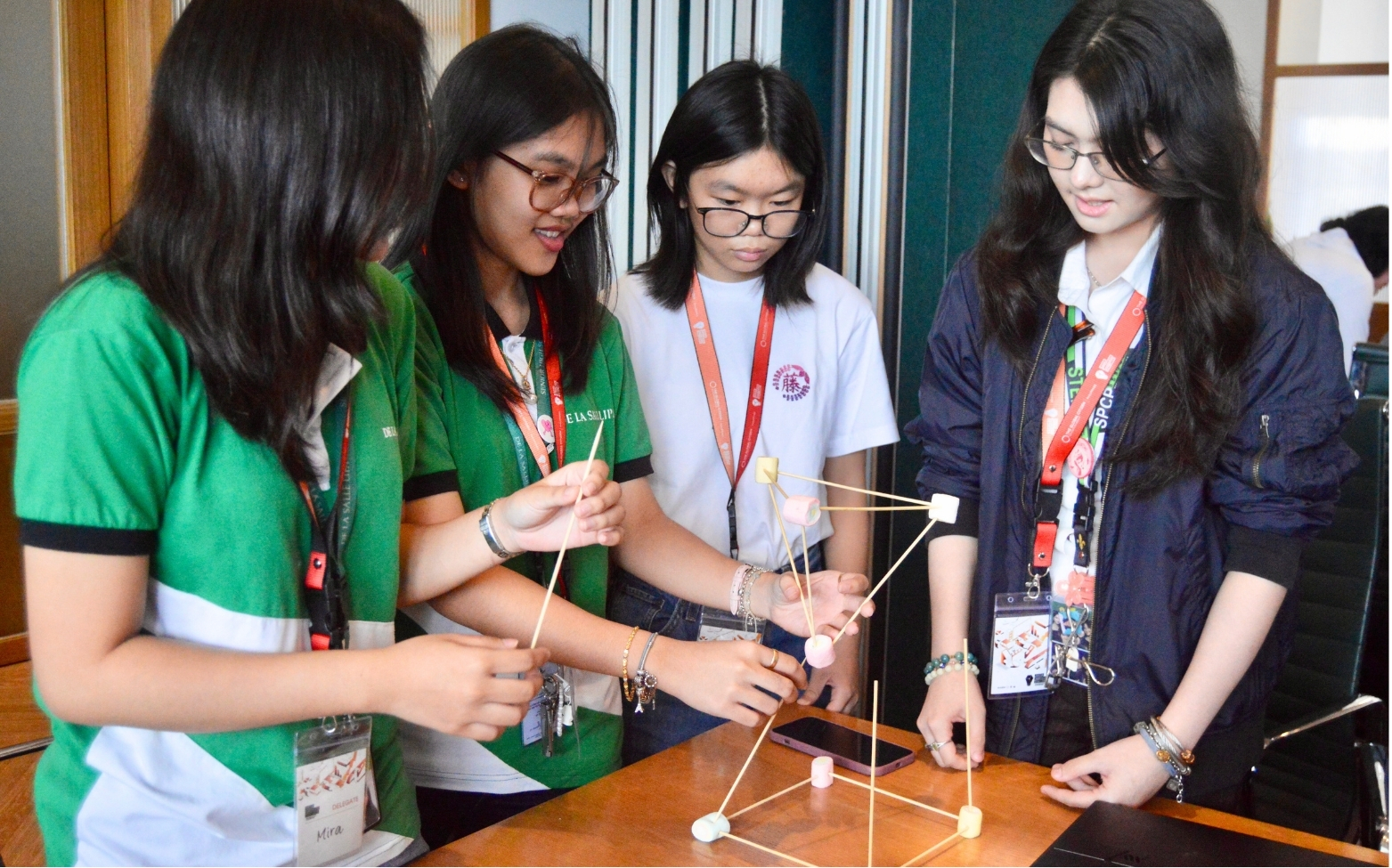 Four young women collaborate to build a geometric structure using marshmallows and wooden sticks around a wooden table.