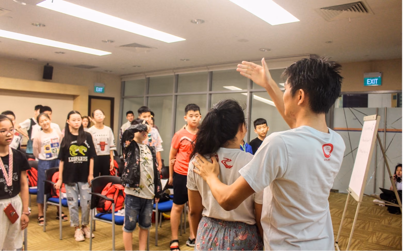 Instructor guiding a group of students standing in a classroom with chairs and a whiteboard.