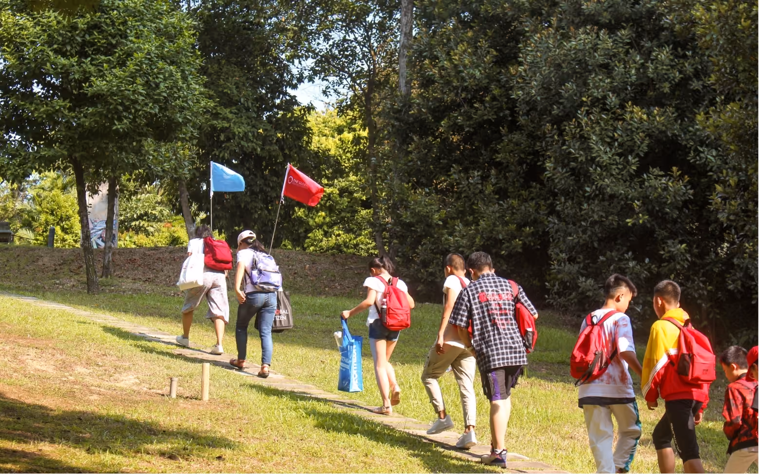 Group of children walking along a narrow path in a green park, carrying backpacks and some holding flags.