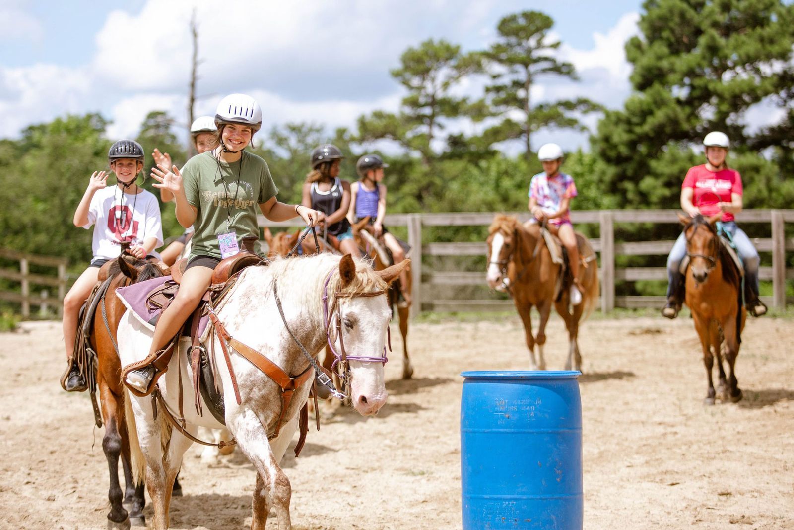 Children wearing helmets riding horses in an outdoor riding arena with a blue barrel in the foreground.