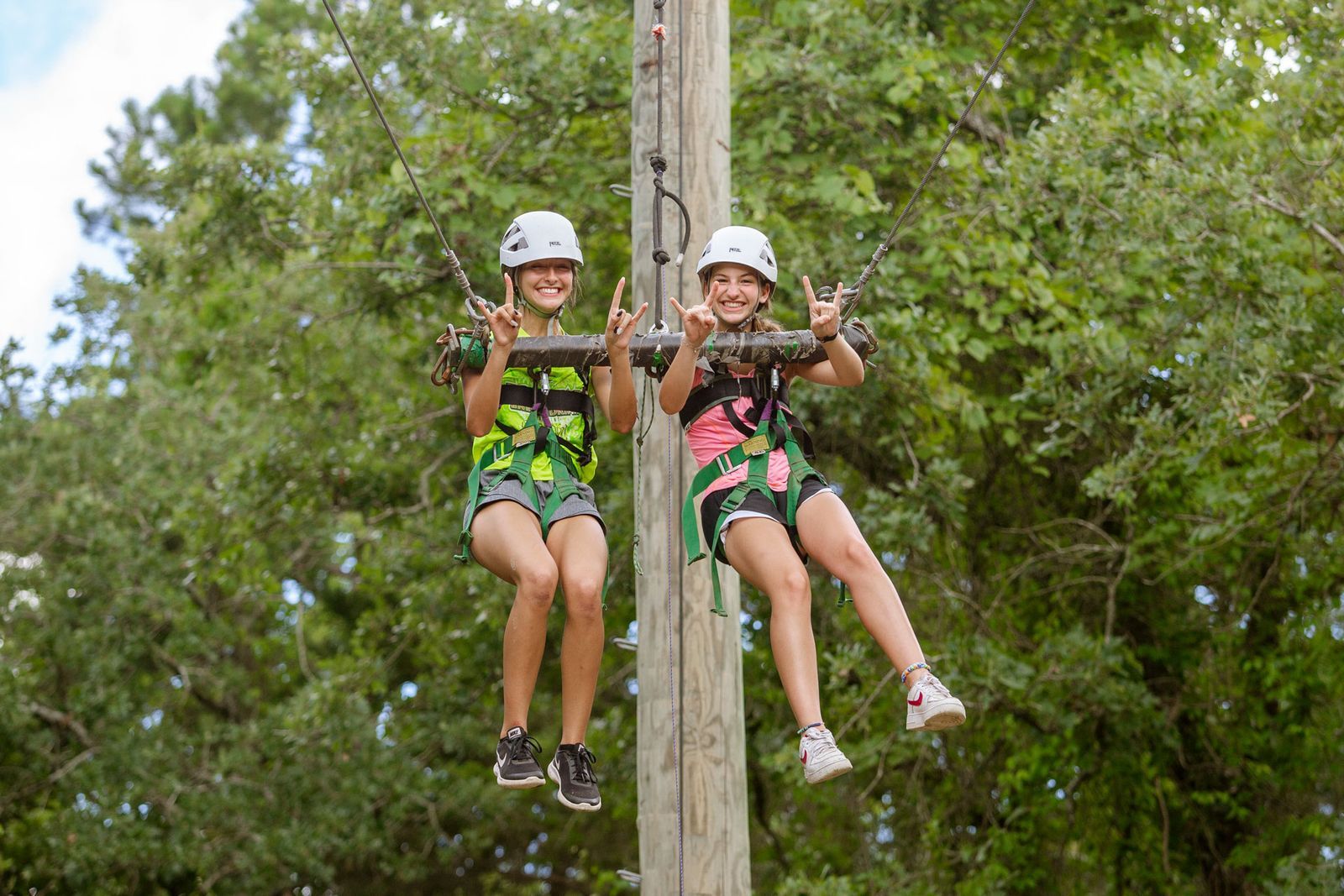 Two girls wearing helmets and safety harnesses smiling and making hand signs while suspended on a ropes course against a background of green trees.