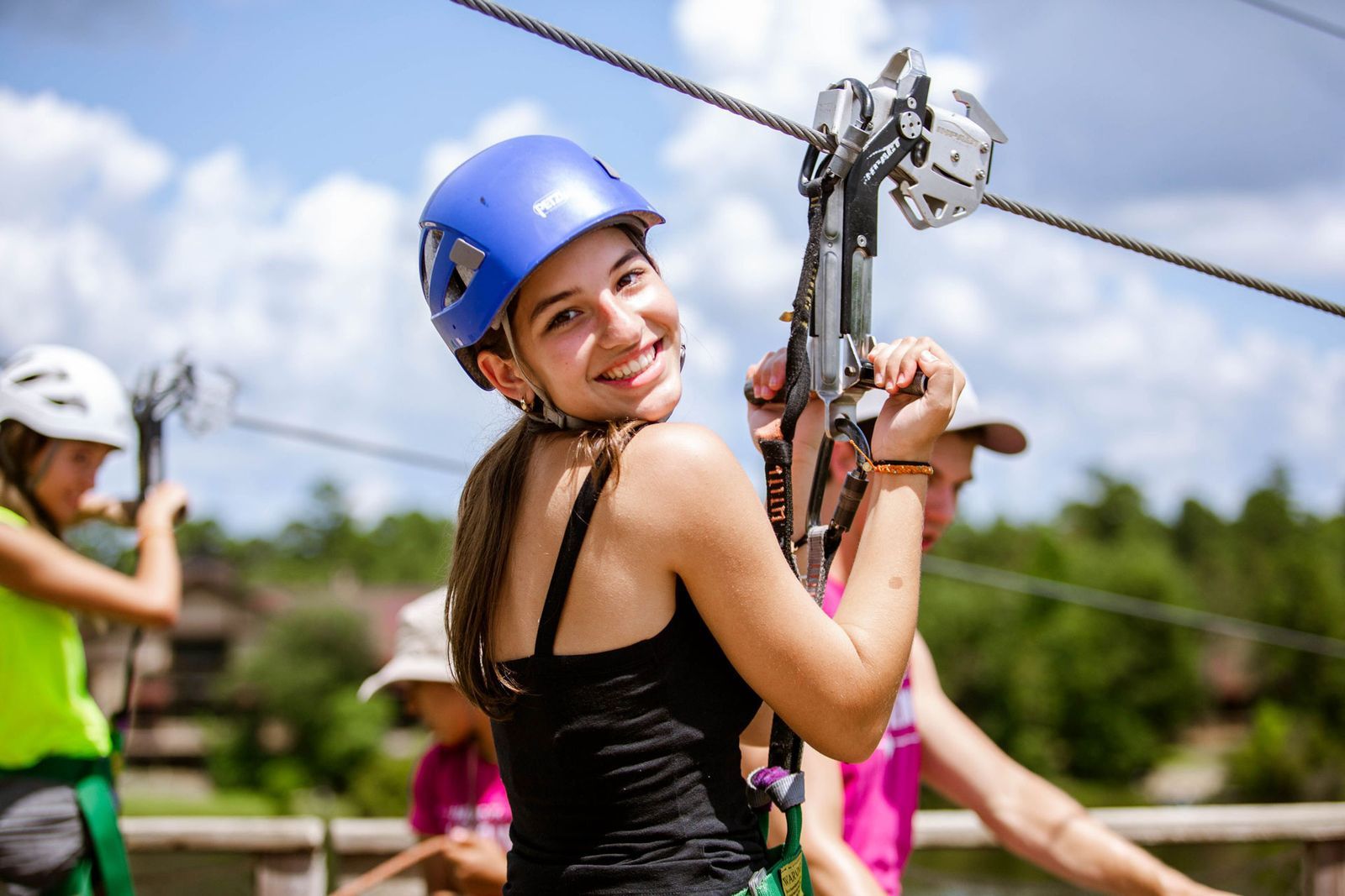 Smiling young girl wearing a blue helmet and black tank top holding a zip line pulley on a sunny day.