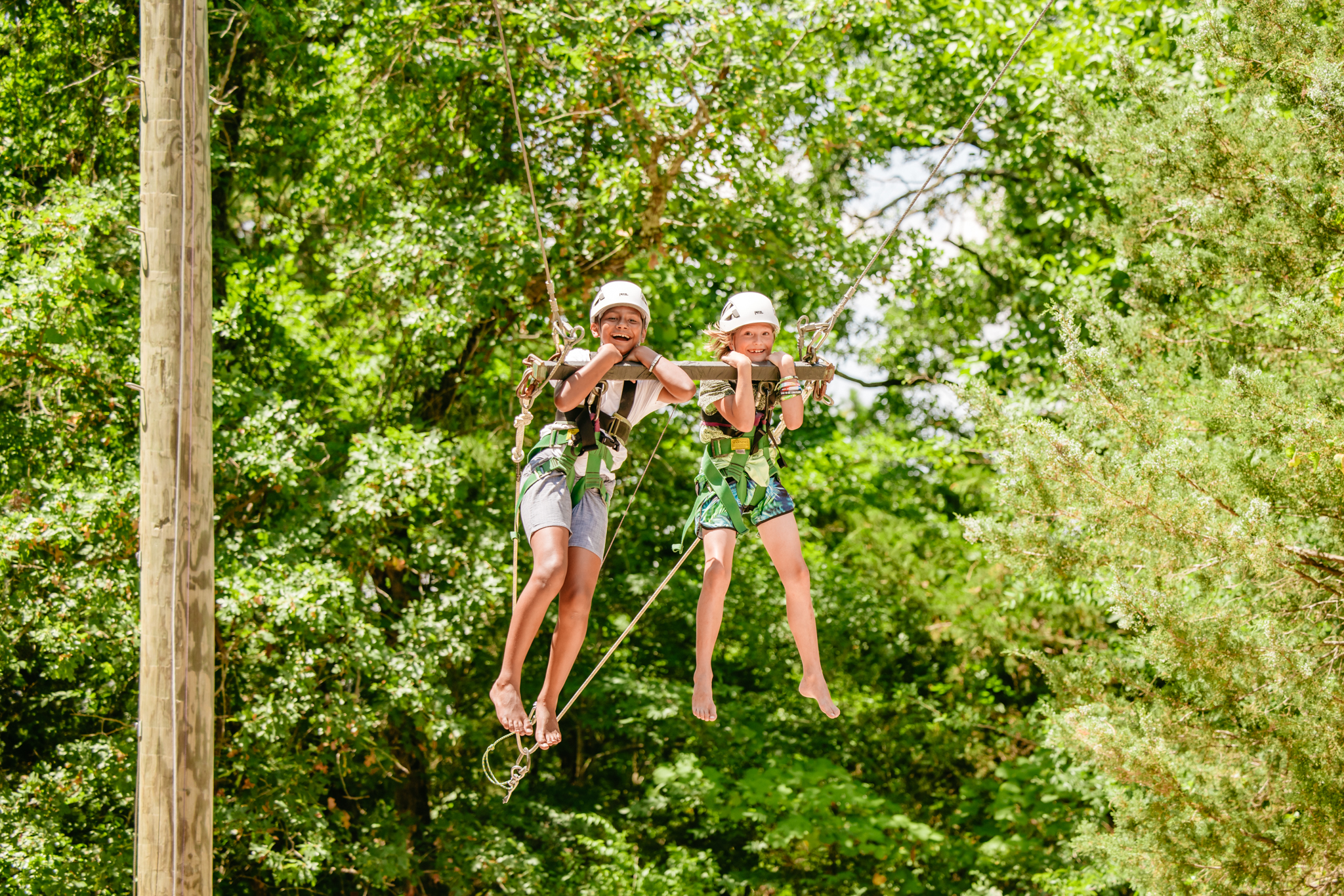 Two children wearing helmets and harnesses smiling while suspended on a zip line swing outdoors surrounded by green trees.