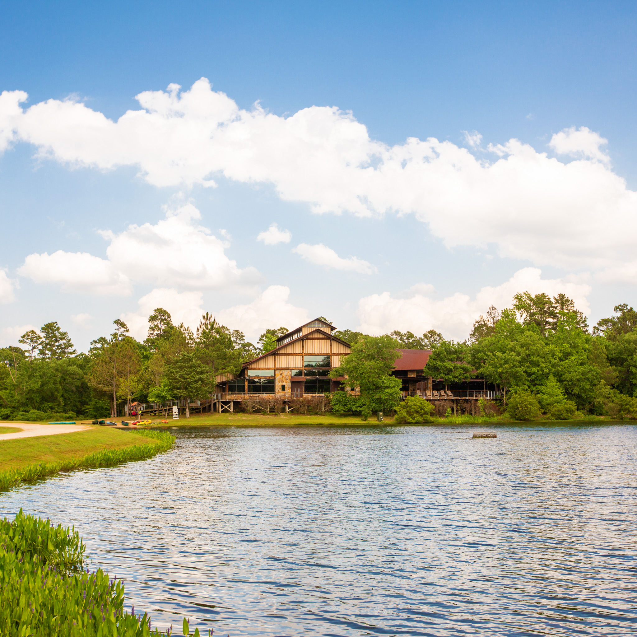 Lakeside view of a rustic lodge surrounded by trees under a blue sky with scattered clouds.