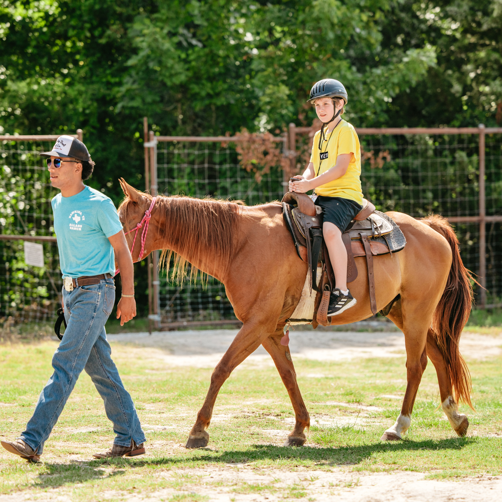 A young boy wearing a helmet rides a brown horse led by a man in a blue shirt and jeans in an outdoor fenced area.