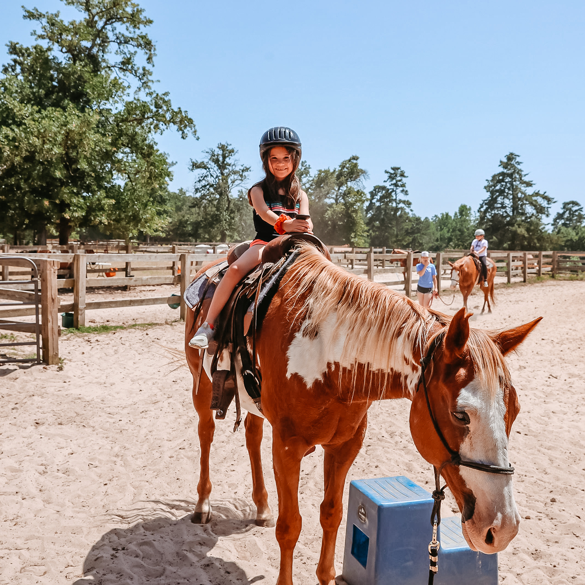 Smiling girl wearing a helmet sitting on a brown and white horse in a sandy paddock on a sunny day.