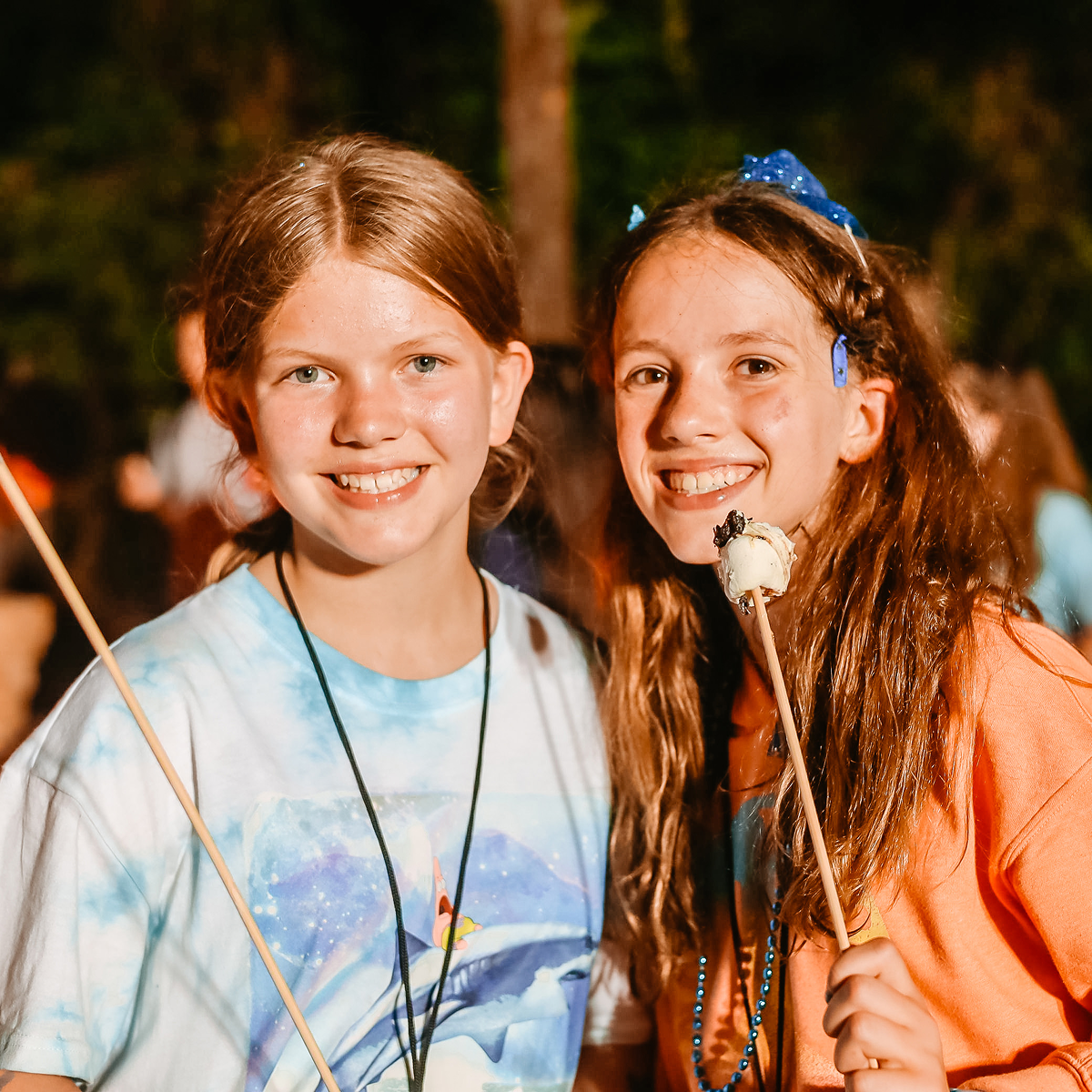Two smiling girls outdoors at night, one holding a stick with a roasted marshmallow.