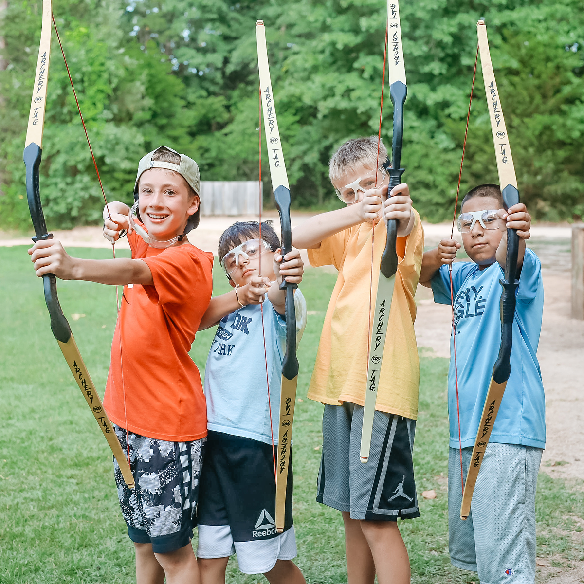 Four boys outdoors practicing archery, aiming bows with safety glasses on.