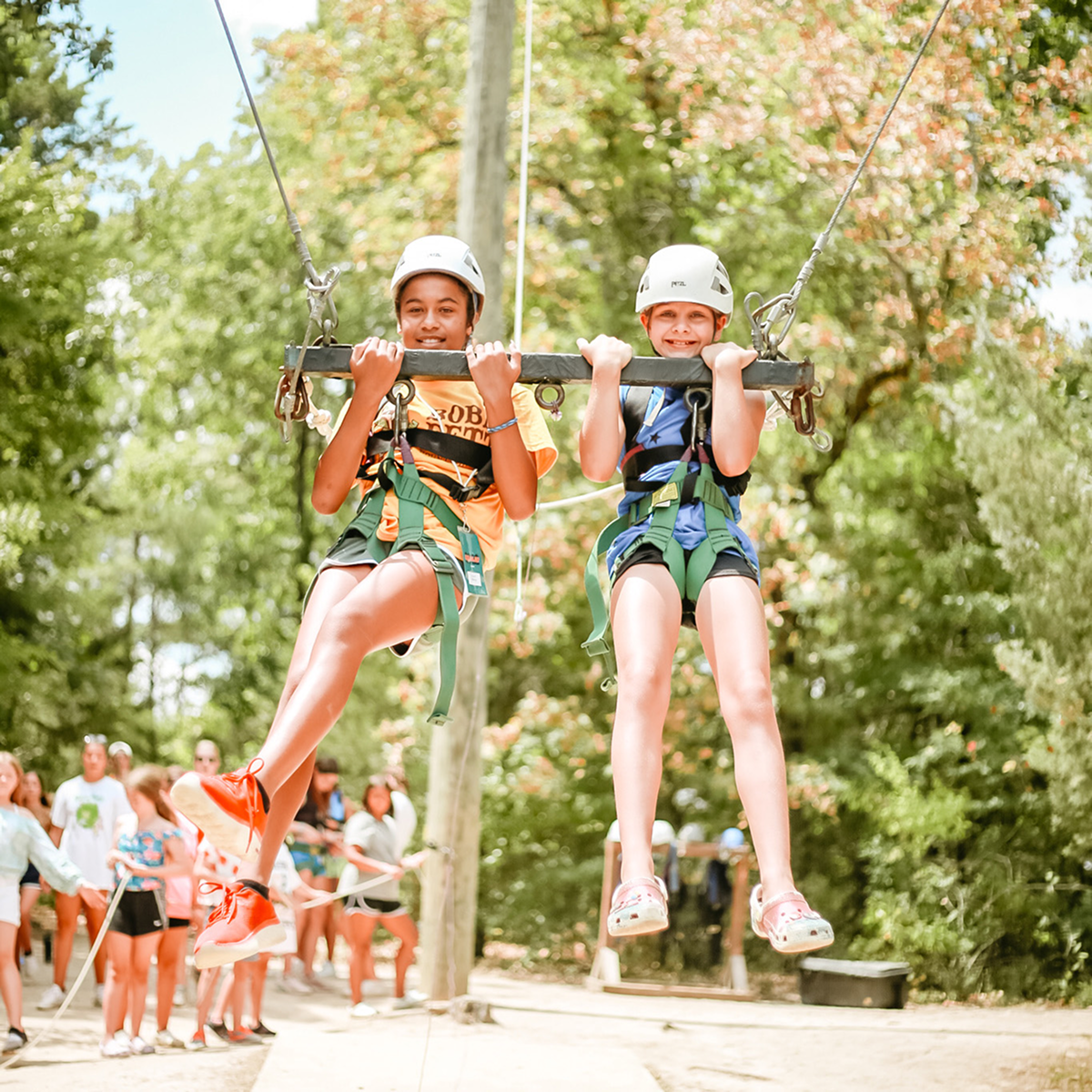 Two children wearing helmets and harnesses on a zipline swing with trees and people in the background.