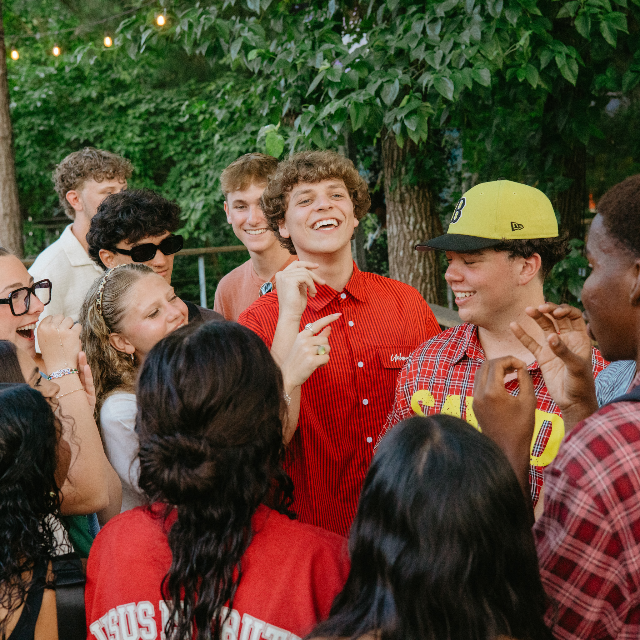 Group of diverse teenagers smiling and laughing together outdoors under string lights and trees.