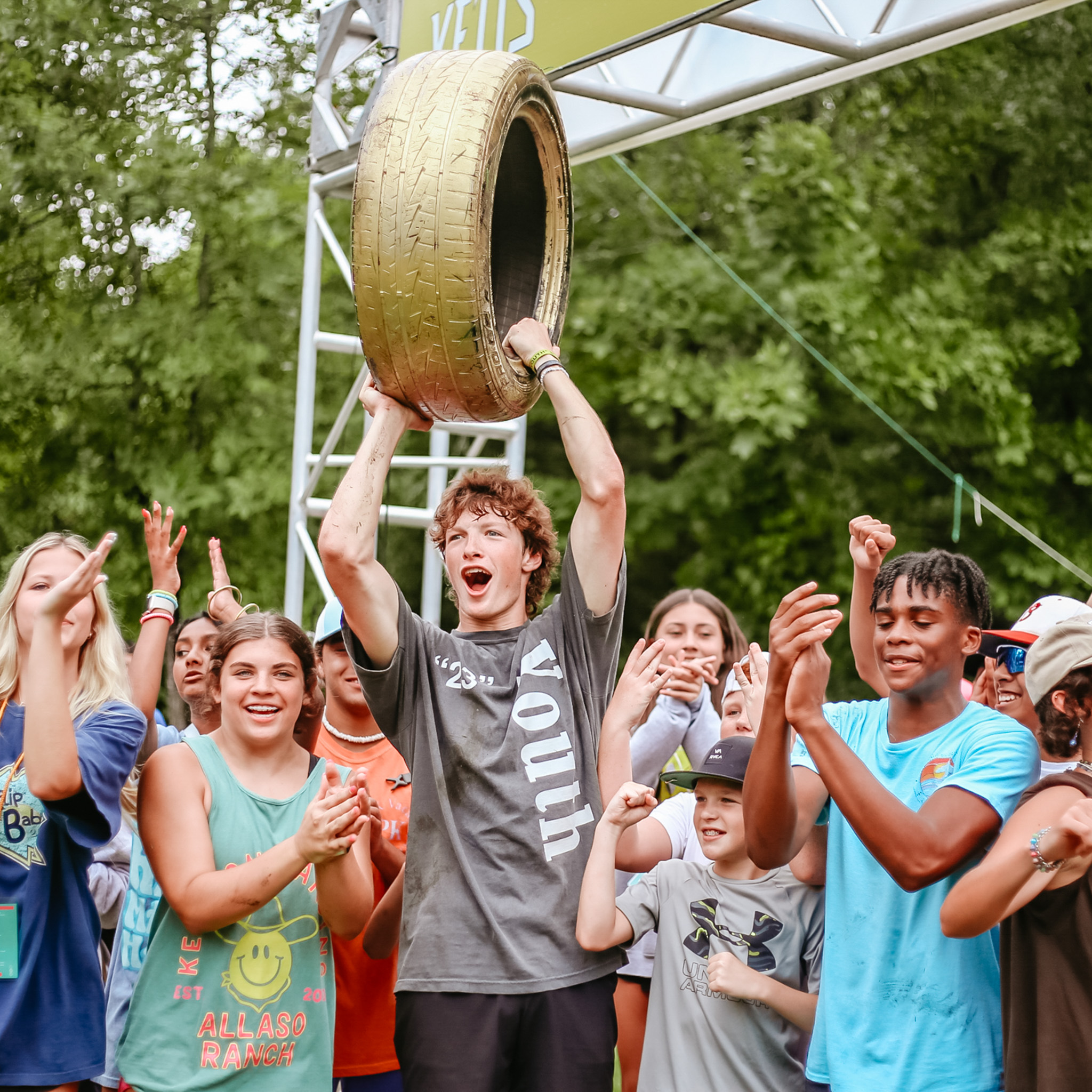 Group of diverse young people celebrating outdoors, with one boy holding a large golden tire trophy above his head.