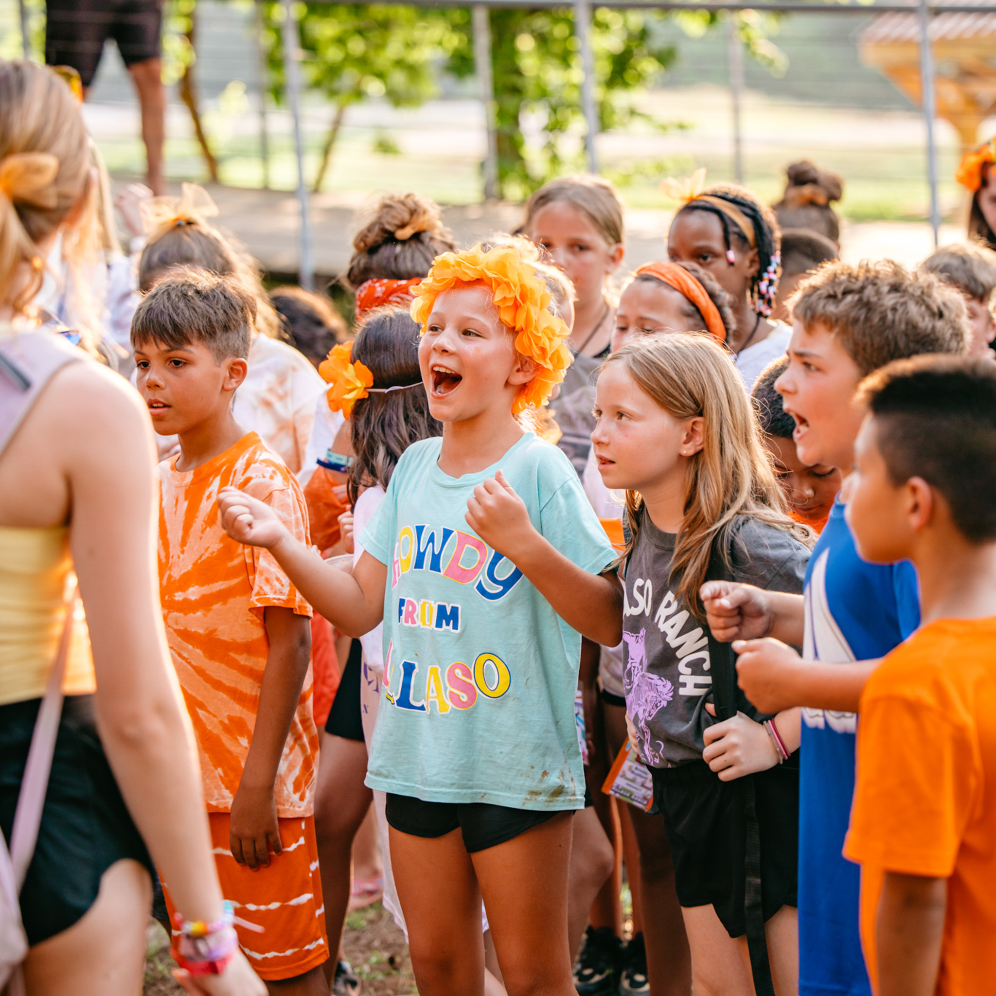 Group of excited children in colorful clothes and floral headbands gathered outdoors, engaging with an adult.