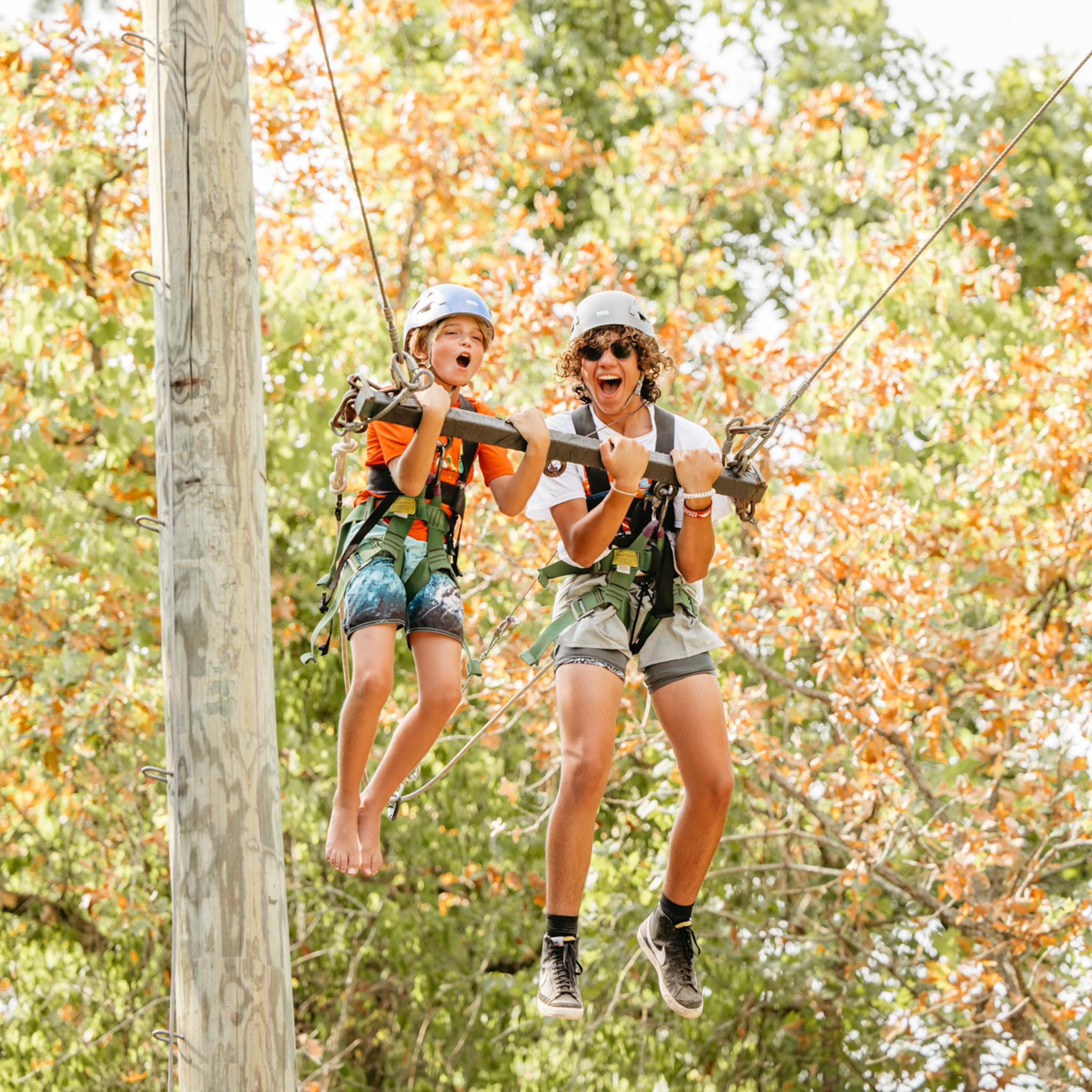Two children wearing helmets and harnesses swinging together on a zip line with green and orange autumn trees in the background.
