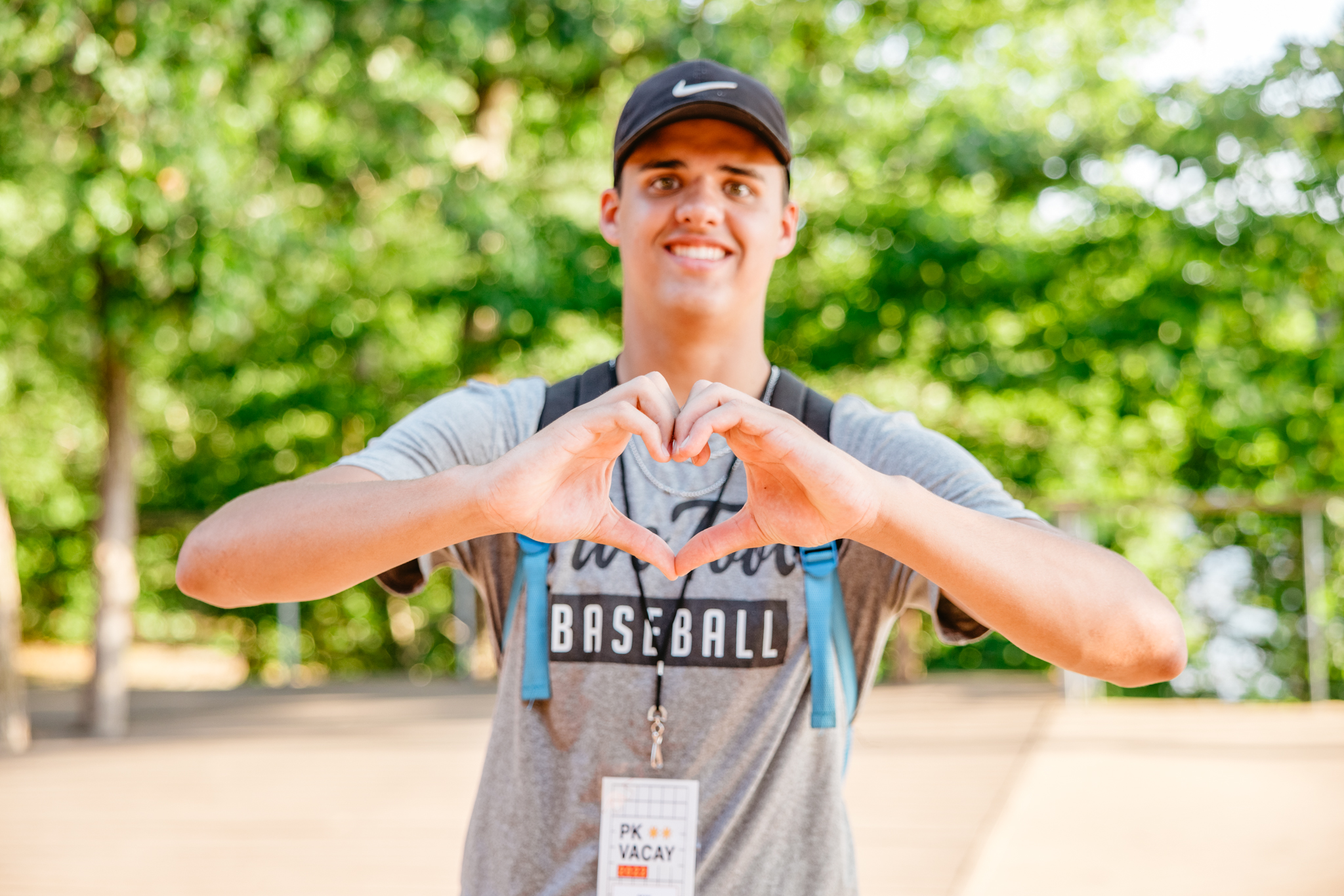 Young man wearing a black cap and gray baseball t-shirt forming a heart shape with his hands outdoors.